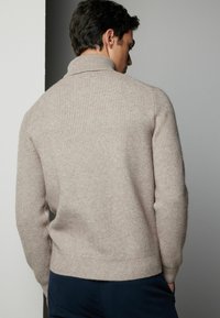 Krämfärgad ribbad tröja med hög krage, texturerad finish och figurnära silhuett, matchad med marinblå byxor, sedd bakifrån.