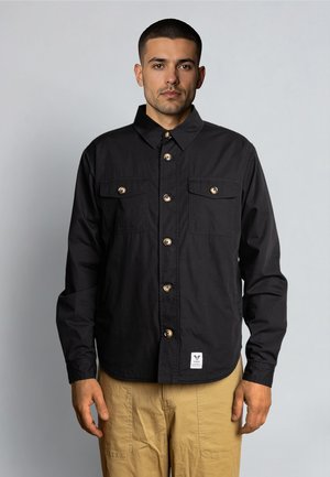 Mand iført sort skjorte med knapper og to brystlommer og beige bukser, stående mod en ensartet lysegrå baggrund.