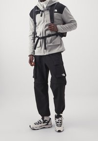 Grau und schwarze Zip-Hoodie mit Fronttaschen, kombiniert mit schwarzen Cargo-Hosen und schwarz-weißen Sneakers. Verfügt über verstellbare Riemen und ein Logo.