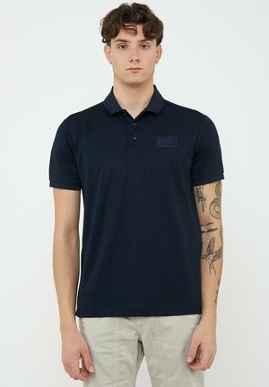 Giovane uomo con tatuaggi sul braccio destro che indossa una polo blu scuro e pantaloni beige chiaro su uno sfondo bianco semplice.