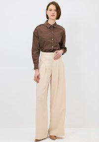 Camicia marrone con colletto strutturato; pantaloni beige a vita alta e a gamba larga; tessuto morbido; design minimale; scarpe tan a punta.