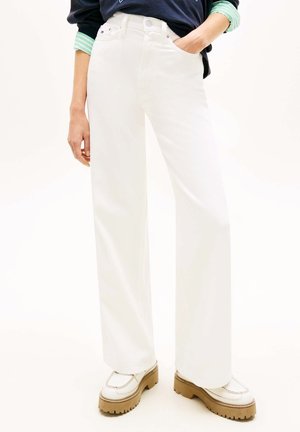 Persona che indossa pantaloni bianchi a vita alta e gamba larga, con mano in tasca e scarpe platform bianche con suole marroni.