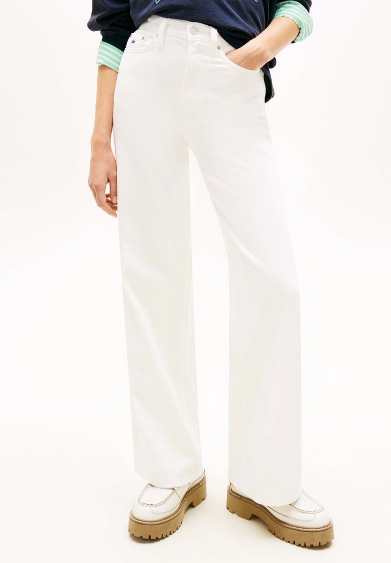 Personne portant un pantalon blanc taille haute à jambes larges, la main dans la poche, et des chaussures plateforme blanches avec des semelles marron.