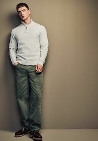Veste zippée gris clair en tissu texturé, associée à un pantalon vert ample et des chaussures lacées marron foncé avec une semelle crantée.