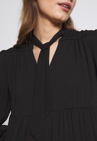 Blouse noire avec un détail de col à nouer, manches longues, texture plissée et décolleté en V. Fabriquée en tissu léger.