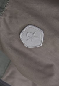 Tissu gris avec un motif en chevrons, présentant un patch hexagonal embossé avec un logo. La couture est visible le long des bords.
