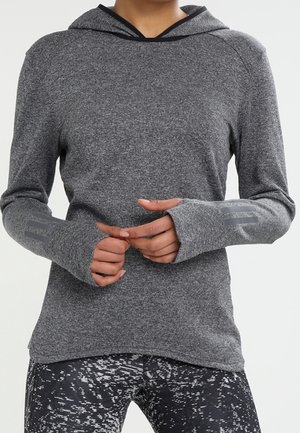Person trägt ein graues Kapuzenoberteil mit langen Ärmeln und Daumenlöchern sowie schwarze Leggings mit Muster, die Hände locker vor der Taille verschränkt.