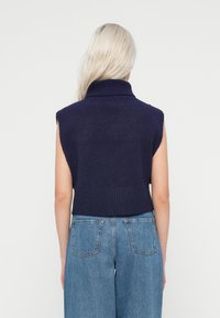 Camisola de malha sem mangas em azul marinho com gola alta e bainha e ombros canelados, combinada com calças de ganga de perna larga em azul claro. Textura suave no geral.