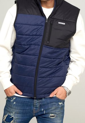 Mann trägt schwarze und marineblaue gesteppte Weste mit Reißverschluss über weißem Sweatshirt, die Hände in den Taschen von abgenutzten blauen Jeans.