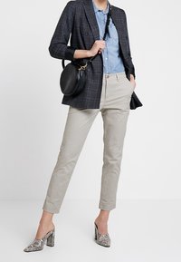 Femme portant un pantalon beige, une chemise bleue, un blazer à carreaux sombre, des talons blocs au motif serpent, et portant un sac à bandoulière noir rond.