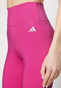 Legginsy wysokiego stanu w intensywnym różowym kolorze, z gładką teksturą i białym logotypem na lewej udzie. Wzmocnione detale szwów.