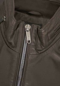 Braune Lederjacke mit hohem Kragen, silbernem Mittelreißverschluss, seitlichen Taschenreißverschlüssen und glatter Textur, die durch Steppnähte akzentuiert wird.