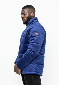 Marinblå vadderad jacka i mjukt tyg, med hög krage, dragkedjestängning och sidopatch med logotyp. Slät yta och funktionell design.