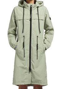 Veste longue à capuche vert clair avec fermeture éclair à l'avant, cordons noirs, poignets à boutons et poches latérales portée par une personne les mains dans les poches.