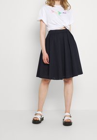 Svart A-linjeformad kjol med texturerat mönster, tillsammans med en vit knyttee grafisk t-shirt och vita sandaler med svarta sulor. Enkelt design.