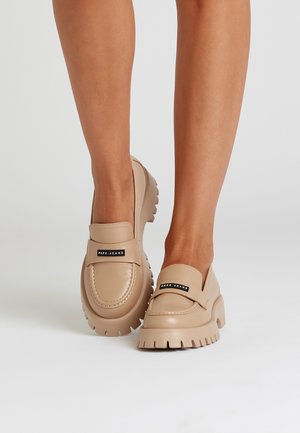 Legs wearing beige chunky-soled loafers with "Pepe Jeans" logo plaques on the straps against a plain light background.