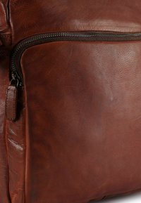 Borsa in pelle marrone con una texture liscia, dotata di una tasca con cerniera e una leggera lucentezza, che evidenziano il materiale resistente e la maestria artigianale.