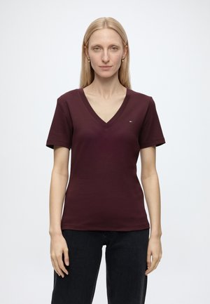 Vrouw met steil blond haar, gekleed in een donkerpaars V-hals T-shirt en zwarte broek, staand tegen een effen lichte achtergrond.