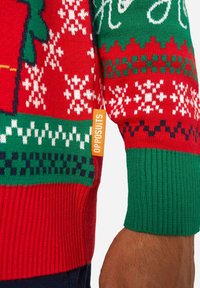 Raudonas ir žalias megztinis su snaigių, raštų motyvais ir etiketė "OPPOSUITS". Ribotos rankogaliai ir apačia, tekstūruota medžiaga.