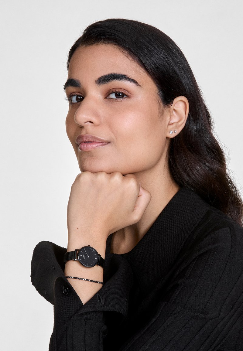 Montre noire avec un cadran rond et un bracelet en maillage noir, portée au poignet. Le porteur a une peau claire et des cheveux foncés, vêtu d'un haut noir.