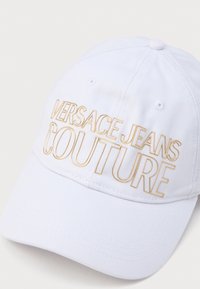 Fehér baseball sapka arany hímzett "VERSACE JEANS COUTURE" felirattal az elülső részen, ívelt karimával és texturált anyaggal.