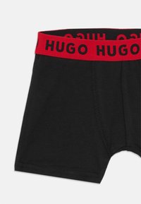 Čierne bavlnené boxerky s červeným elastickým pásom, na ktorom je v čiernom písmene napísané "HUGO". Obsahujú ploché švy a majú priliehavý strih.
