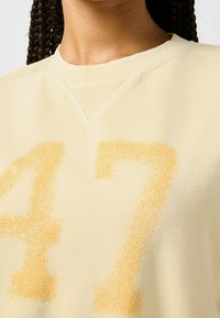 Nahaufnahme eines hellgelben Sweatshirts mit der verblassten Nummer 47 in einem dunkleren Gelb, getragen von einer Person mit geflochtenem Haar.