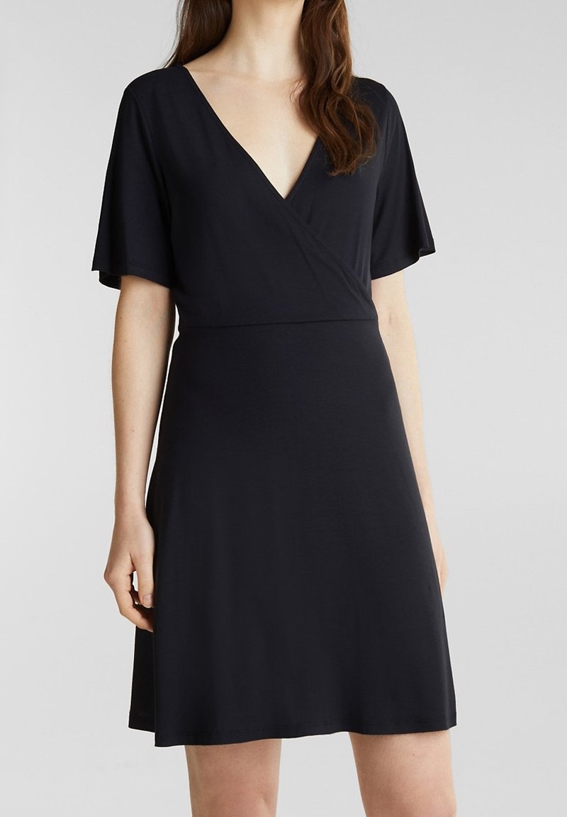 Femme portant une robe portefeuille noire longueur genou avec des manches courtes et un décolleté en V, sur un fond gris clair uni.