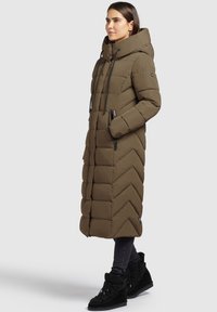 Olvai zaļš pūkains mētelis ar garu, ševronveida dizainu, lielu kapuci, auklītēm un ar rāvējslēdzējiem aizveramām kabatām. Valkāts ar tumšām biksēm un melnajām kurpēm.