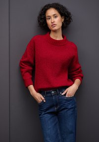 Rode gebreide trui met een hoge kraag, ontspannen pasvorm en omgerolde mouwen, gecombineerd met donkerblauwe jeans en een gladde textuur.