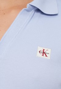 Lys blå ribbestrikket poloskjorte med oppbrettet krage, med en rektangulær Calvin Klein-logo i rødt og hvitt.