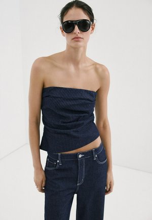 Mujer con gafas de sol negras de gran tamaño, top azul marino con rayas de espiguilla sin tirantes y vaqueros a juego con rayas de espiguilla, sobre un fondo blanco liso.