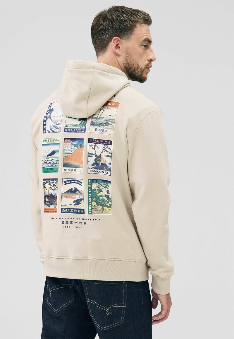 Beige hoodie med et trykt grafisk motiv på ryggen, der viser forskellige landskaber og historiske scener arrangeret i et gittermønster.