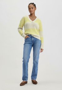 V-Ausschnitt-Pullover in gelb-creamfarbenen Streifen, strukturiertes Strickmuster. Kombiniert mit blauen Denim-Jeans und braunen flachen Schuhen, posiert das Modell stehend.