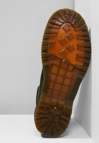 Suela de calzado negra con un patrón de banda de rodadura robusto y hendiduras profundas; textura de goma con acentos transparentes en marrón.