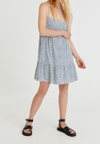 Ärmelloses Kleid mit blauem Blumenmuster, gestuftem Rock und dünnen Trägern, kombiniert mit schwarzen Sandalen mit Riemchen. Glatte Stofftextur.