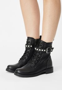 Černé kožené kotníkové boots s krajkovým zapínáním, cvokovými akcenty, páskem s dekorativními perlami a texturovaným bočním panelem.