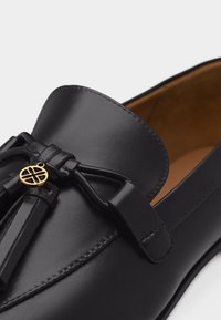 Μαύρο δερμάτινο μοκασίνι με φούντες και μικρό χρυσό έμβλημα στο μπροστινό μέρος, εσωτερική επένδυση σε καφέ χρώμα, κοντινή άποψη.