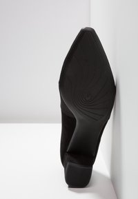 Chaussure à talon haut en daim noir avec une semelle en caoutchouc texturée, présentant un design effilé et des lignes courbes subtiles, inclinée sur un fond blanc.