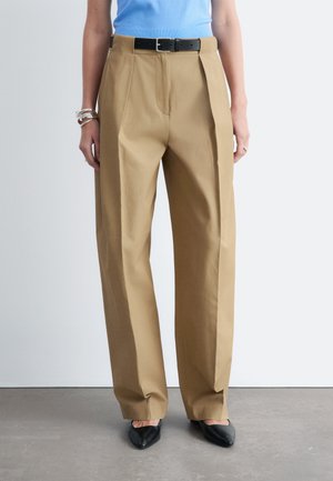 Personne portant un pantalon beige à jambes larges avec une ceinture noire, un haut bleu sans manches, des bracelets argentés et des chaussures pointues noires, debout sur un sol gris.