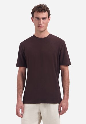 Man with curly hair wearing a plain dark brown short-sleeve t-shirt and light beige pants, standing against a white background.