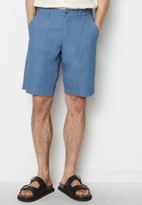 Mężczyzna w niebieskich szortach do kolan i czarnych klapkach z srebrnymi klamrami, stojący przy gładkiej białej ścianie.