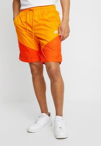 Plavky Nike s barevným blokovým designem v oranžové a žluté barvě, elastickým páskem a bočními kapsami. Materiál je lehký a hladký.