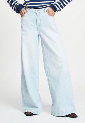 Lichtblauwe jeans met wijde pijpen, gedragen met handen in de zakken en schoenen met luipaardprint, gecombineerd met een marineblauw-wit gestreept top met lange mouwen.