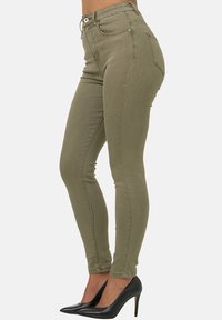Jeans skinnies verde oliva con cintura alta, bolsillos frontales y detalles cosidos, combinados con zapatos de tacón negro.