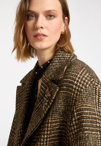 Brązowy płaszcz w kratę houndstooth z teksturowanej wełny, z klapami, w oversize'owym kroju, z kolorową broszką jako akcentem.