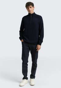 Marineblauer Strickpullover mit hohem Kragen und Halbreißverschluss, kombiniert mit dunklen Jeans und weißen Sneakers. Glatte Textur, klassischer Schnitt.