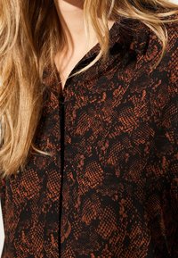 Schwarze Bluse mit einem strukturierten Rostmuster, mit einem kragenähnlichen Ausschnitt und Knopfdetails, die einen weichen Stoffausführung zeigt.