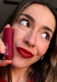 Röd läppstiftstub som hålls bredvid ansiktet, med en matt finish. Modellen har djupröda läppar och bruna ögon, med vågigt hår och hoopörhängen.