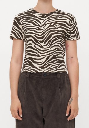 Person iført en kortærmet top med zebra print og højtaljede mørkebrune manchesterbukser, med synlige tatoveringer på begge underarme.